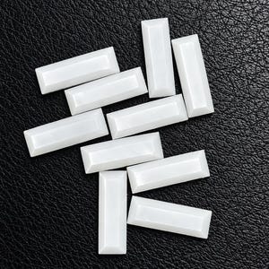 Puede incluir: Un conjunto de nueve cabujones rectangulares blancos con bordes biselados y un acabado liso y brillante. Estas piezas pequeñas y alargadas están dispuestas en un patrón disperso, mostrando su forma uniforme y su color blanco opaco.