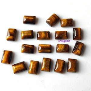 May include: A collection of rectangular tiger's eye gemstones. The polished stones display a mix of golden brown and dark brown hues, with a chatoyant effect. The text "smkgems" is visible on one of the stones.