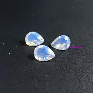 Peut inclure: Trois pierres précieuses opalescentes en forme de poire, avec une irisation blanche laiteuse et bleue. Les pierres sont présentées sur une surface sombre. Les pierres sont probablement destinées à la fabrication de bijoux.