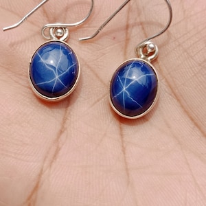 May include: A pair of silver earrings featuring blue star sapphire gemstones. The earrings have a simple design with a small oval-shaped stone suspended from a hook.