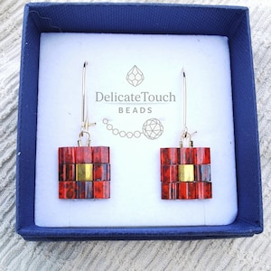 Può includere: Orecchini pendenti quadrati con perline rosse e dorate, presentati in una scatola regalo blu. Gli orecchini hanno un design rettangolare con un centro dorato. Il testo "Delicate Touch BEADS" è visibile.