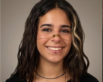 May include: A gold-toned metal nose bridge chain featuring a central flat rectangular plate that rests across the bridge of the nose, connected to thin delicate chains that extend outward toward the cheeks, creating a decorative facial accessory.