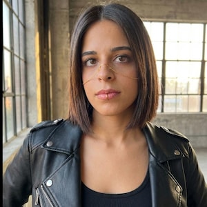 May include: A gold-colored nose and lip chain. The jewelry is worn on the face, with a chain across the bridge of the nose and a lip chain. The person is wearing a black leather jacket and a black top.