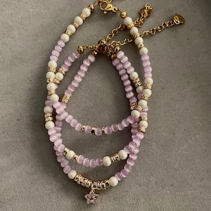 Op de afbeelding: Drie gelaagde armbanden met kralen. De armbanden hebben een combinatie van witte en lichtpaarse kralen, geaccentueerd met goudkleurige metalen afstandhouders en sluitingen. Eén armband heeft een sterbedel.