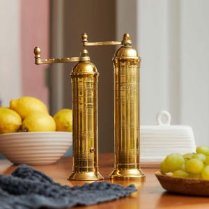 May include: Two gold-colored pepper grinders with a crank handle, standing on a wooden surface. The grinders are cylindrical with a detailed design. A bowl of lemons, a bowl of grapes, and a dark blue cloth are also visible.