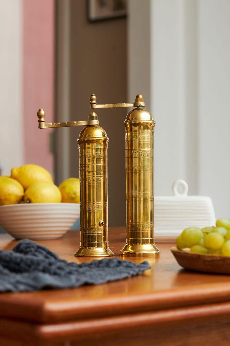 May include: Two gold-colored pepper grinders with a crank handle, standing on a wooden surface. The grinders are cylindrical with a detailed design. A bowl of lemons, a bowl of grapes, and a dark blue cloth are also visible.
