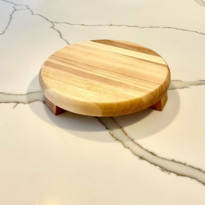 May include: A round wooden cake stand with a light-colored top and a darker wood base. The stand is approximately 8 inches in diameter and is elevated by three small, rectangular feet. The wood grain is visible, adding to its natural aesthetic.
