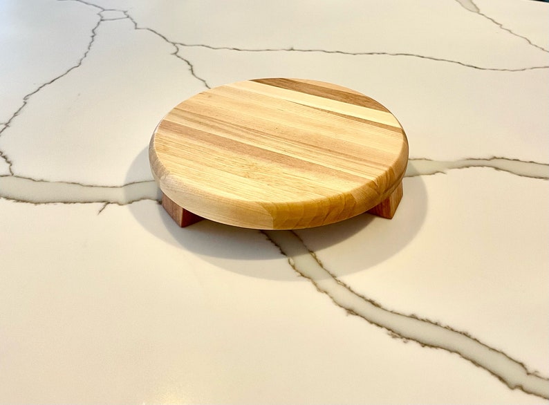 May include: A round wooden cake stand with a light-colored top and a darker wood base. The stand is approximately 8 inches in diameter and is elevated by three small, rectangular feet. The wood grain is visible, adding to its natural aesthetic.