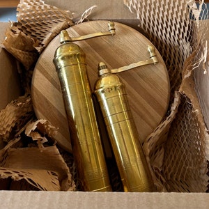 May include: Two brass pepper grinders with a wooden cutting board inside a cardboard box. The grinders are cylindrical with a crank handle and decorative etched details. The cutting board is round with a light wood grain.