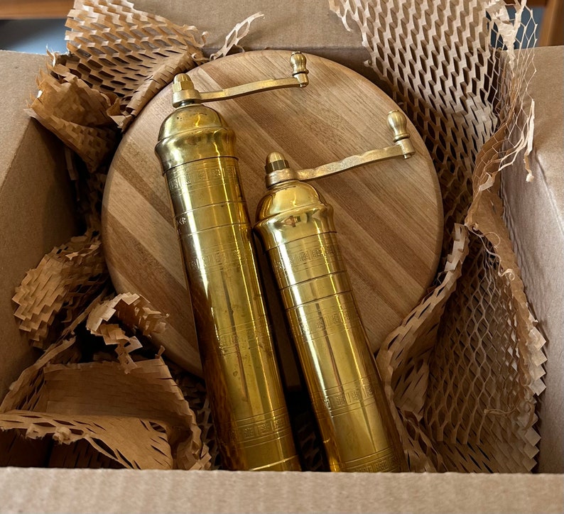 May include: Two brass pepper grinders with a wooden cutting board inside a cardboard box. The grinders are cylindrical with a crank handle and decorative etched details. The cutting board is round with a light wood grain.