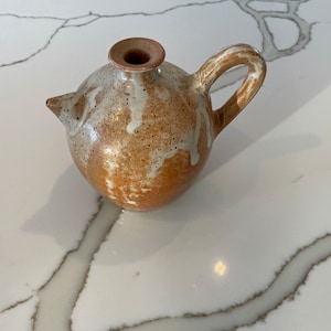 May include: A brown and white ceramic pitcher with a speckled glaze and a rounded body. The pitcher has a single handle and a spout.