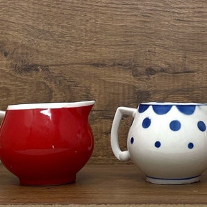 May include: Three ceramic creamer pitchers, one red, one white with blue polka dots, and one red. The pitchers are on a wood surface.