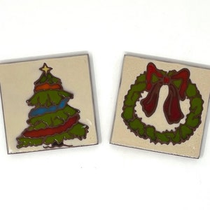 May include: Two ceramic tiles with Christmas designs. One tile features a green Christmas tree with a yellow star on top. The other tile features a green wreath with a red bow.