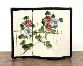 May include: A white ceramic tile with a black border, featuring a hand-painted floral design of pink and blue flowers with green leaves. The tile has a wavy, uneven edge.