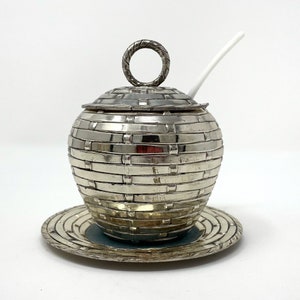 May include: Silver sugar bowl with a woven design and a matching saucer. The bowl has a lid with a ring handle and a small white spoon.