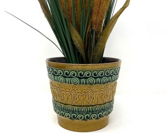 May include: A brown and green ceramic planter with a decorative design. The planter has a wide mouth and a narrow base. It is filled with a green plant with brown stalks.