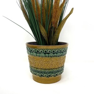 May include: A brown and green ceramic planter with a decorative design. The planter has a wide mouth and a narrow base. It is filled with a green plant with brown stalks.