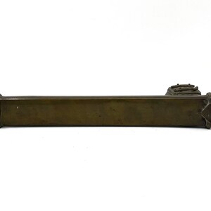 May include: A long, rectangular, bronze-colored metal object with a decorative design on the end. The object is smooth and has a slightly aged appearance.