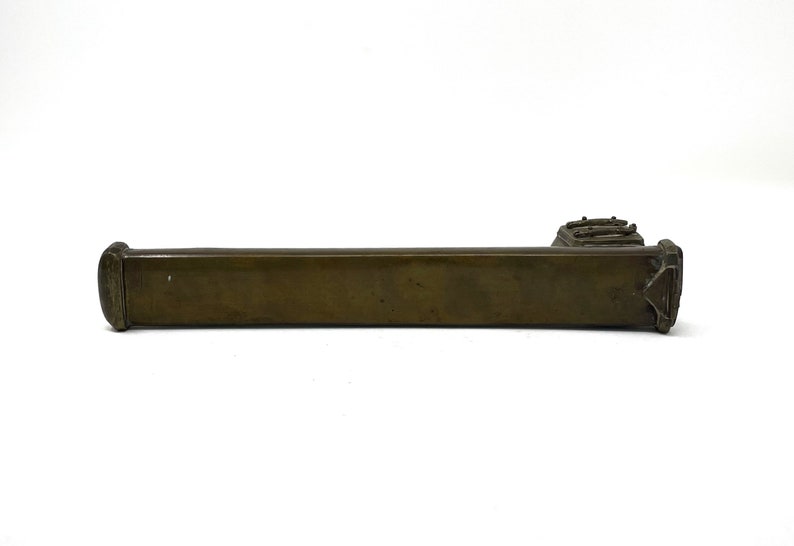 May include: A long, rectangular, bronze-colored metal object with a decorative design on the end. The object is smooth and has a slightly aged appearance.