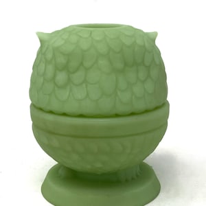 May include: A light green owl-shaped container with a lid. The container has a textured, scale-like pattern on the owl's head and body. The base is round with a decorative pattern. The owl has small, pointed ears and is made of a solid, opaque material.