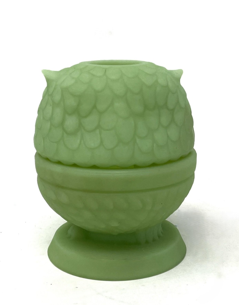 May include: A light green owl-shaped container with a lid. The container has a textured, scale-like pattern on the owl's head and body. The base is round with a decorative pattern. The owl has small, pointed ears and is made of a solid, opaque material.