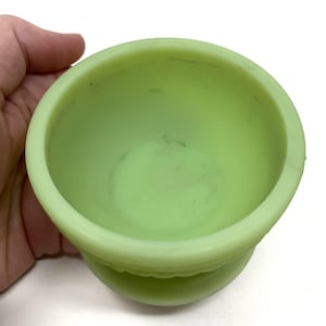 May include: A light green, small, round bowl with a pedestal base. The bowl has a smooth, matte finish and a slightly textured base. The interior of the bowl shows some dark marks. The bowl is held by a hand.