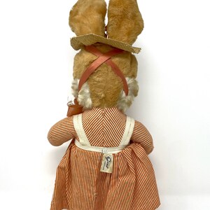 Op de afbeelding: Een pluche bruin konijn met een strooien hoed en een oranje-wit gestreepte jurk. Het konijn houdt een kleine oranje emmer vast. Het konijn heeft een label op zijn jurk met de tekst "Elsie Creation".