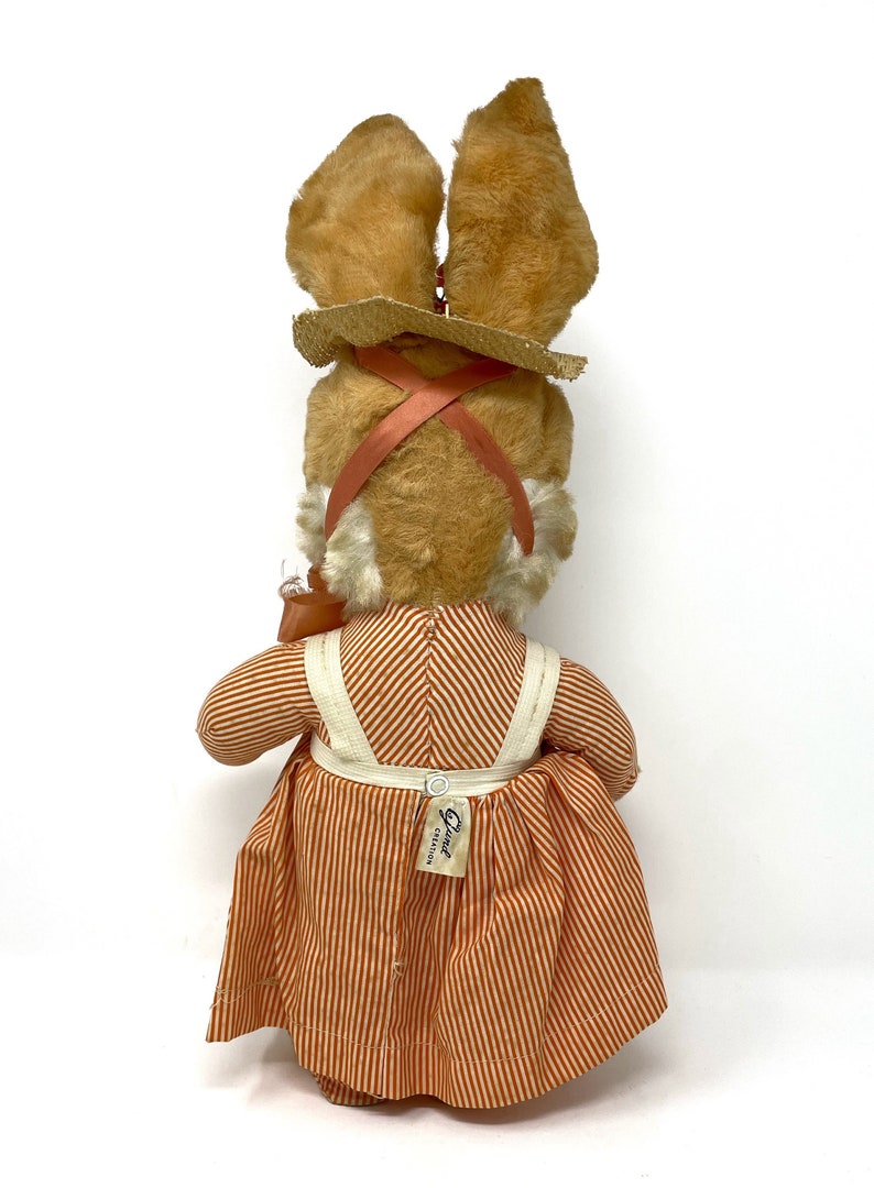 Op de afbeelding: Een pluche bruin konijn met een strooien hoed en een oranje-wit gestreepte jurk. Het konijn houdt een kleine oranje emmer vast. Het konijn heeft een label op zijn jurk met de tekst "Elsie Creation".