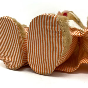 Op de afbeelding: Een pluche speelgoed met oranje en wit gestreept stof. Het speelgoed ligt op zijn rug met zijn voeten in de lucht. Het speelgoed heeft een harige kop en een witte kraag.