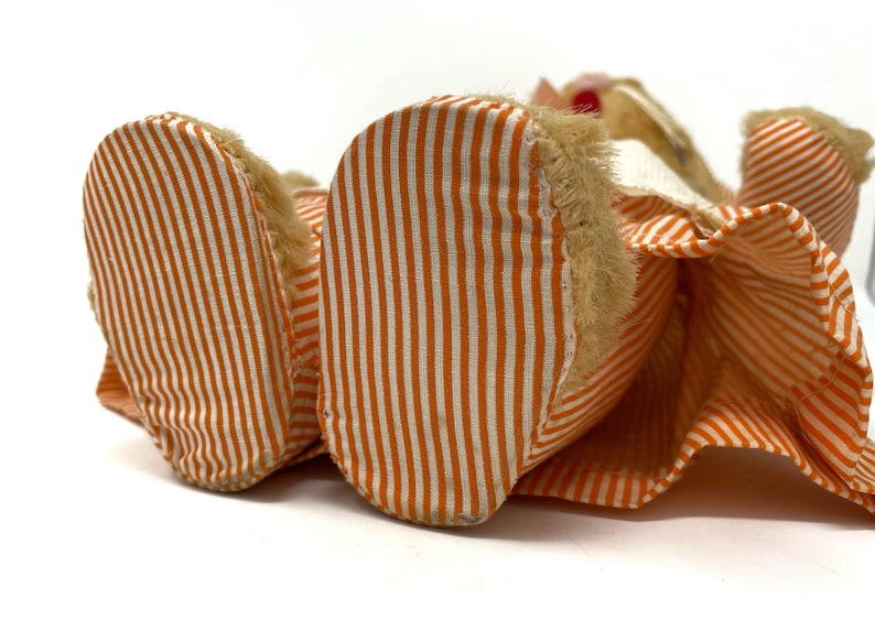 Op de afbeelding: Een pluche speelgoed met oranje en wit gestreept stof. Het speelgoed ligt op zijn rug met zijn voeten in de lucht. Het speelgoed heeft een harige kop en een witte kraag.