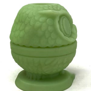 May include: A light green owl-shaped decorative container. The container is made of a solid material and features a detailed design of feathers and an owl's face. The container is round and sits on a small, round base.