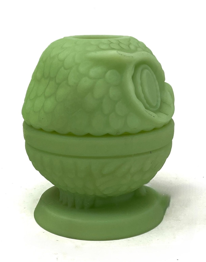 May include: A light green owl-shaped decorative container. The container is made of a solid material and features a detailed design of feathers and an owl's face. The container is round and sits on a small, round base.