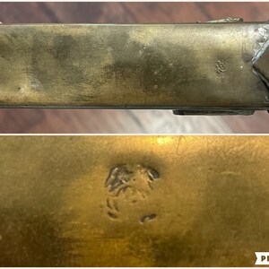 May include: A close-up of a brass, rectangular box with a decorative design on the top. The box has a small, engraved mark on the bottom.