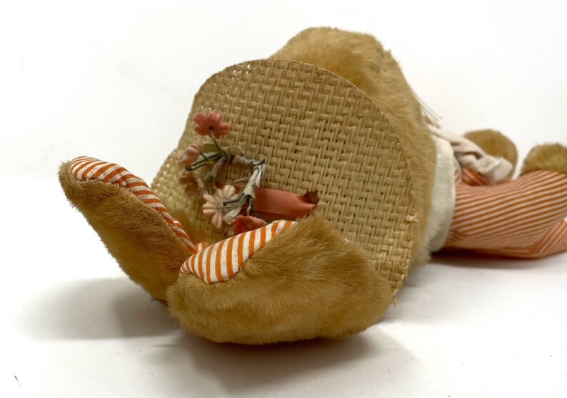 Op de afbeelding: Een pluche konijn speelgoed met een strohoed en oranje-wit gestreepte broek. Het konijn ligt op zijn zij met zijn hoofd op de hoed. De hoed heeft een roze lint en een bloemversiering.