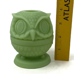 May include: A light green owl-shaped decorative object. The owl has large oval eyes and a textured surface. The object is approximately 4 inches tall and sits on a round base. A yellow ruler is next to the owl.