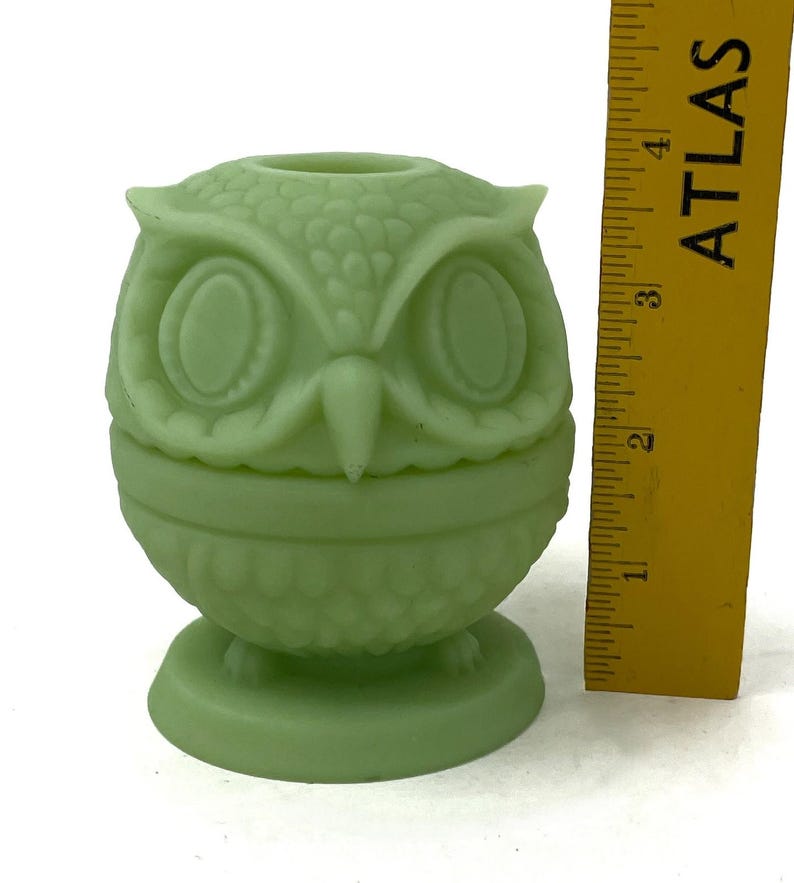 May include: A light green owl-shaped decorative object. The owl has large oval eyes and a textured surface. The object is approximately 4 inches tall and sits on a round base. A yellow ruler is next to the owl.