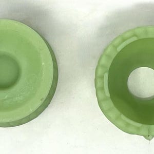 May include: Two light green glass Fenton candle holders. One has a Fenton sticker. The candle holders are round with a central hole. One has a decorative edge. The candle holders are likely vintage.