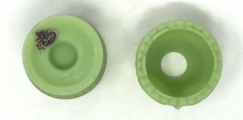 May include: Two light green glass Fenton candle holders. One has a Fenton sticker. The candle holders are round with a central hole. One has a decorative edge. The candle holders are likely vintage.