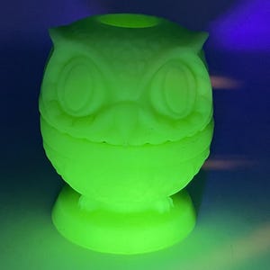 May include: A glowing green owl-shaped figurine. The figurine has large, round eyes, a beak, and ear tufts. It is sitting on a round base. The owl is illuminated with a bright green light, creating a striking visual effect.