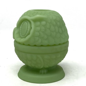 May include: A light green owl-shaped trinket box. The box is made of glass and has a detailed design of an owl's feathers and facial features. The trinket box is approximately 4 inches tall.