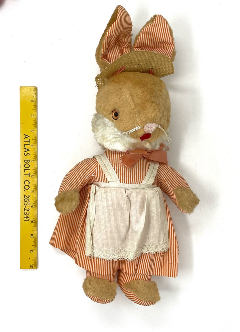 Op de afbeelding: Een vintage pluche konijnen speelgoed dat een strohoed en een jurk met oranje en witte strepen draagt. Het konijn heeft een roze strik en een witte schort met kant.