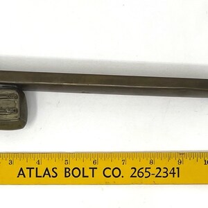 May include: A brass-colored metal rod with a square end and a square-shaped base. The rod is about 10 inches long. The base has a rectangular opening with a raised edge. The rod is sitting on a yellow ruler with the text "ATLAS BOLT CO. 265-2341".