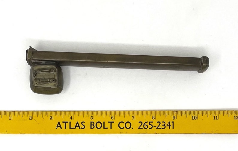 May include: A brass-colored metal rod with a square end and a square-shaped base. The rod is about 10 inches long. The base has a rectangular opening with a raised edge. The rod is sitting on a yellow ruler with the text "ATLAS BOLT CO. 265-2341".