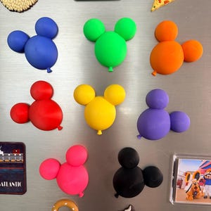 May include: A collection of colourful Mickey Mouse-shaped balloon magnets in blue, green, orange, red, yellow, purple, pink, and black. Additional magnets include a chocolate-dipped treat, a pizza slice, a rectangular sign, and a character photo.