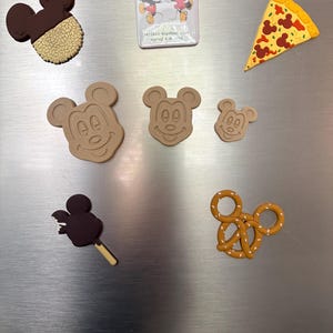 May include: A collection of seven decorative magnets including a chocolate-dipped cookie, a square magnet with Mickey and Minnie Mouse, a pizza slice, three Mickey Mouse head shapes in varying sizes, a chocolate popsicle, and a pretzel.
