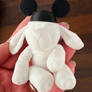 May include: A small, white, 3D-printed figurine of a dog with long, floppy ears. The dog is wearing a black cap with two round, black ears attached to the top. The surface of the white dog shows visible horizontal layer lines from the printing process.