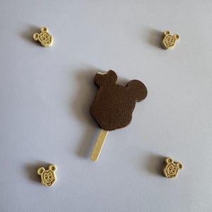May include: A brown chocolate-covered ice cream bar shaped like a mouse head on a wooden stick with a small bite taken out of the top, surrounded by four small, light-coloured, mouse-shaped charms arranged in a rectangular formation.
