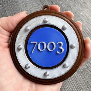 May include: A circular brown, white, and blue ornament featuring the number 7003 in raised white text. The design includes a brown outer ring, a white middle ring with six small silver-coloured studs, and a solid blue centre circle.