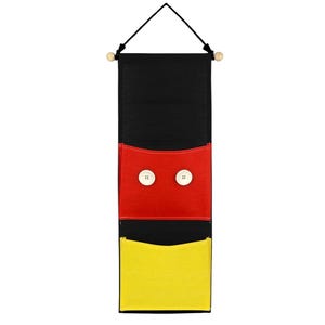 May include: A vertical black fabric wall organiser featuring two pockets. The top pocket is red with two large white buttons attached to the front. The bottom pocket is yellow. The organiser hangs from a wooden dowel with a black cord.