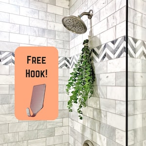 May include: A white tiled shower with a silver shower head and a silver hook. The hook is labeled "FREE HOOK!" and has a green plant hanging from it.